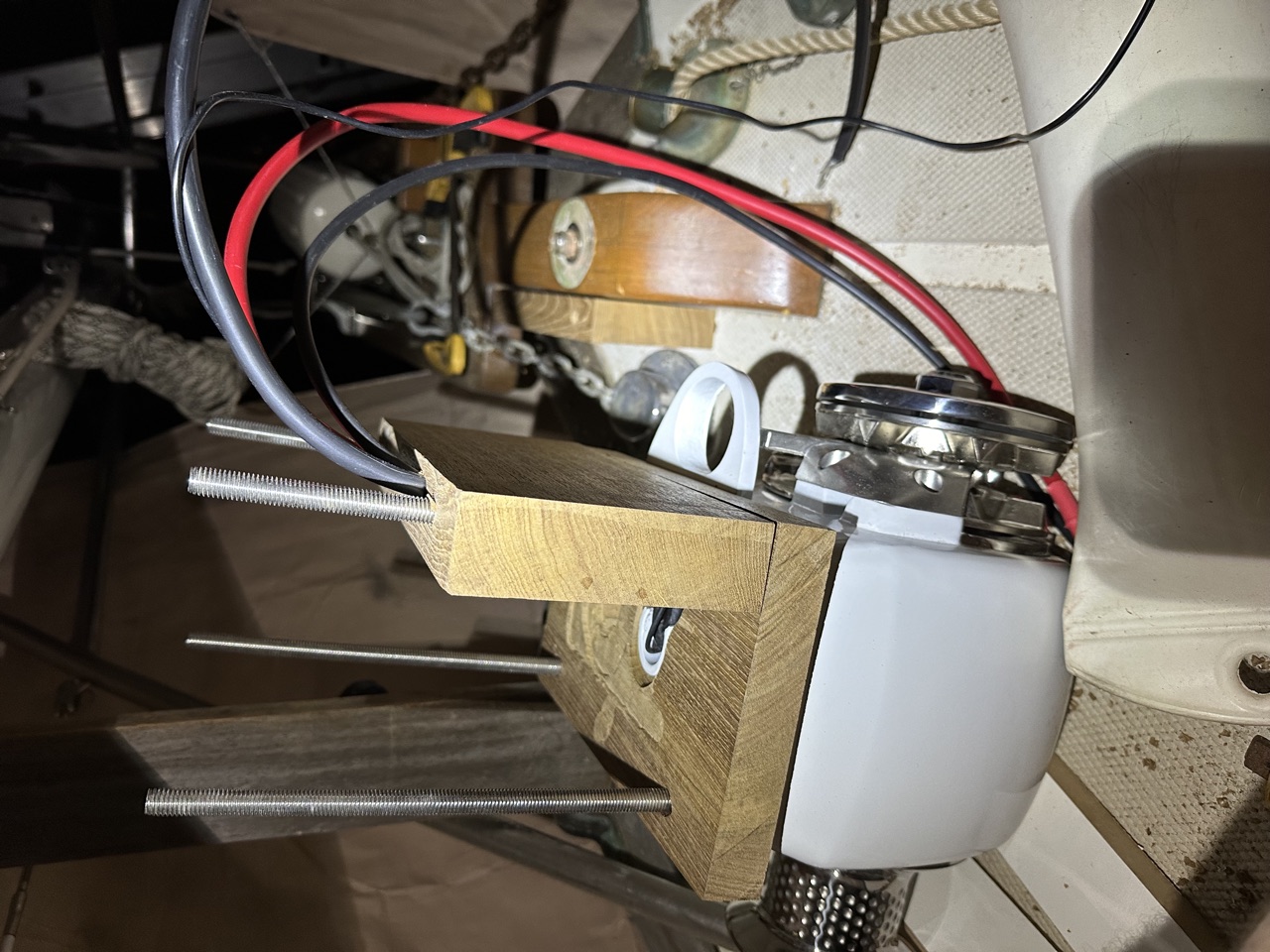 Underside of the bowsprit showing red and black power cables routed through the teak support block with threaded mounting rods protruding